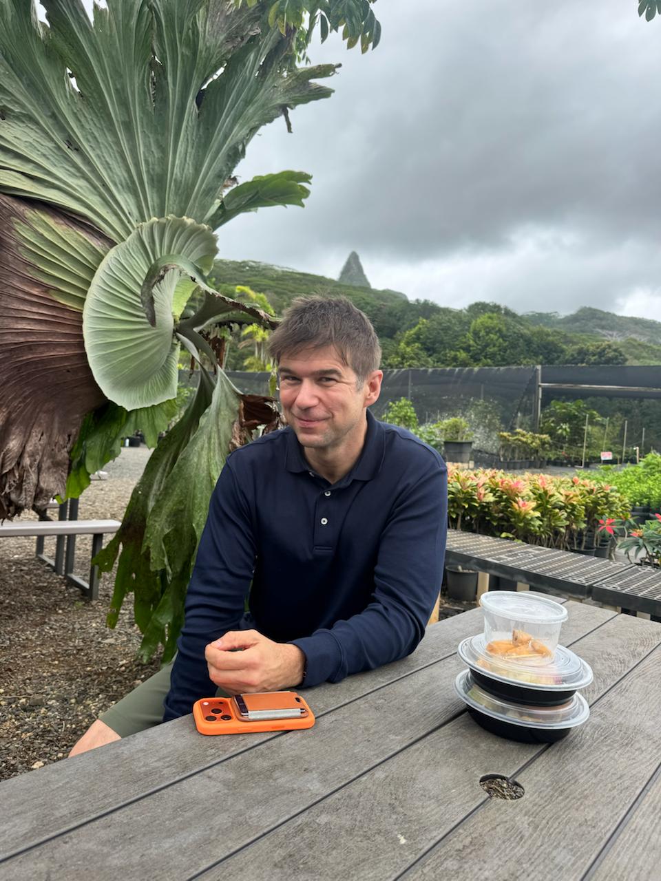 Clay Collins sitting at a picnic table at an outdoor plant nursery, phone on the table in front of him